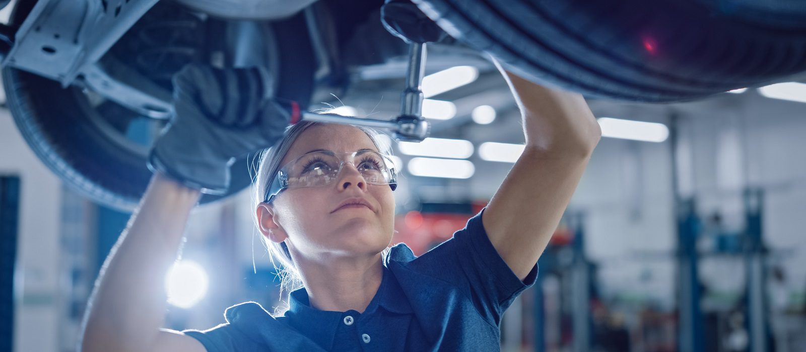 Portrait Shot of a Female Mechanic Working Under Vehicle in a Car Service. Empowering Woman Wearing Gloves and Using a Ratchet Underneath the Car. Modern Clean Workshop.