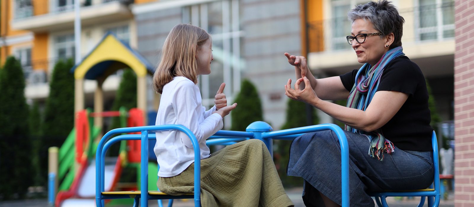 Two people have fun on a blue merry-go-round. They are sitting on the seats and holding the handles. They are teacher and student. They are deaf or learning signs.